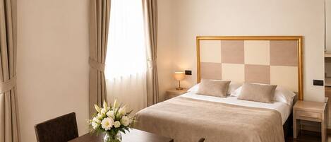 A hotel room with a bed, bedside tables, a desk with chairs, a vase of flowers, and curtains.