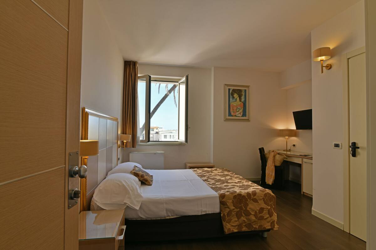 A hotel room with a bed, a desk, a chair, a window, and a painting on the wall.