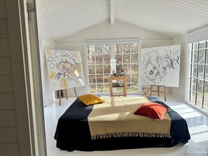 Una habitación con una cama, dos caballetes con pinturas, una ventana y una puerta.