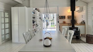 Un espace repas comprenant une longue table, des chaises blanches, un lustre, une bibliothèque et une cuisine avec un fourneau.