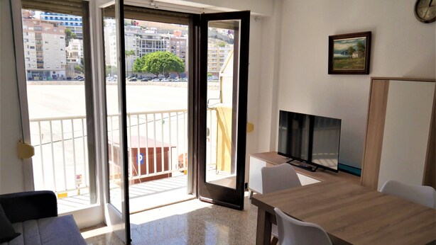 A room with a wooden table, a TV, a sofa, and a balcony with a view of buildings and a tree. - AMANECER, 1º-2 (Cullera)