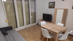 A living room with a grey sofa, a wooden dining table with white chairs, a TV on a stand, and a sliding glass door with blinds. - AMANECER, 1º-2 (Cullera)
