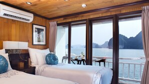 Habitación de hotel con dos camas, una mesita de noche, una mesa con sillas y un amplio ventanal con vistas al mar y a las montañas.