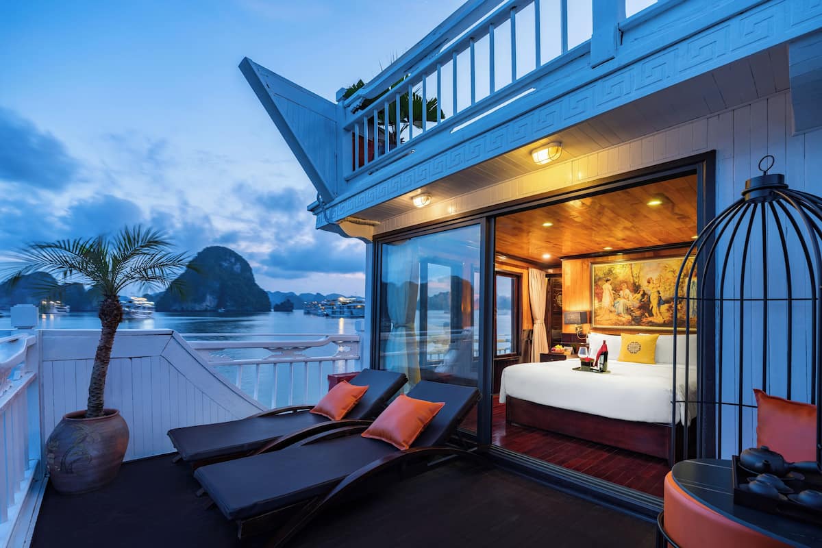 A balcony with sun loungers and a potted plant, a bedroom with a bed and a painting, and a view of boats on water.