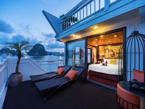 A balcony with sun loungers and a potted plant, a bedroom with a bed and a painting, and a view of boats on water. - Hera Classic Boutique Cruise (Tuan Chau)