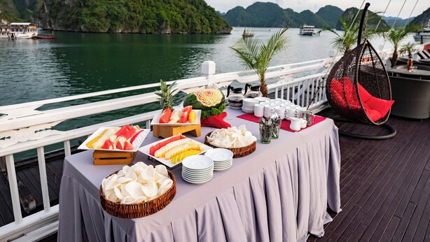 A table set with various fruits and snacks on a boat deck with a view of a mountainous island and the sea. - Hera Classic Boutique Cruise (Tuan Chau)