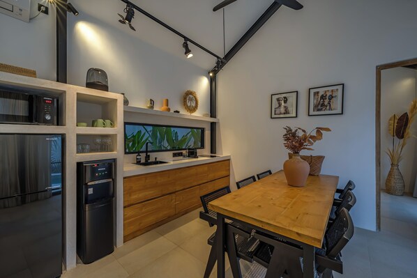 A modern kitchen with a built-in microwave, refrigerator, and coffee maker. There is a wooden dining table with chairs and a large window above it. - Villa Coco.Nuts (Canggu)