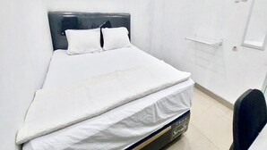Een netjes opgemaakt bed met een zwart hoofdbord en witte beddengoed in een eenvoudige kamer met een witte muur en een klein raam.
