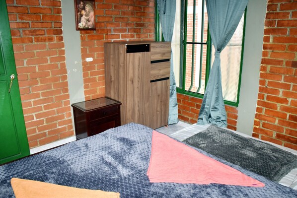 Una habitación con pared de ladrillo, cama con cubrecama estampado, armario de madera, mesita de noche pequeña y ventana con cortinas.