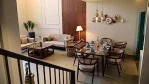 Un comedor con una mesa de madera dispuesta para seis personas, rodeada de sillas de mimbre. En el fondo, un sofá y una mesita con una lámpara son visibles.