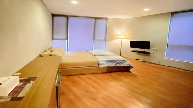 A modern bedroom with a wooden floor, a bed with a striped blanket, a desk with papers, a lamp, and a TV on the wall.
