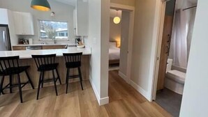 A modern kitchen with a central island, black bar stools, and a view of a bedroom through an open door.