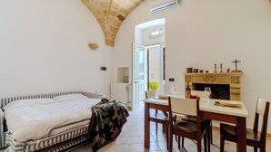 A bedroom with a bed, a dining table with chairs, a fireplace, and a door leading to another room. - Corte dei Mesci - U Farraru - Historic Center Specchia (Specchia)