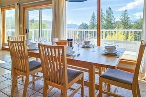 A dining area with a wooden table and chairs, a view of trees and mountains through large windows. - 3 bedroom house with garage (Invermere)