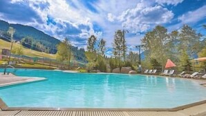 Piscina com espreguiçadeiras e guarda-sóis, cercada por árvores e montanhas.