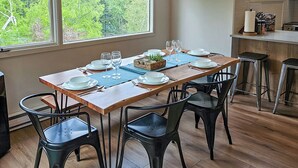Uma sala de jantar com uma mesa de madeira posta para quatro pessoas, rodeada por cadeiras pretas, e grandes janelas com vista para uma floresta.