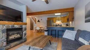 A living room with a stone fireplace, a television, a sofa, and a kitchen area with wooden cabinets and a bar counter. - 3 Bed Townhouse - Fireplace - Great Location (Fernie)
