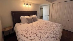 A bedroom with a large bed, a dark headboard, and a wall-mounted light fixture. - 3 BDRM Condo - Superb Location - Parking (Fernie)