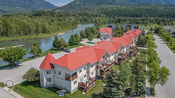 A row of modern, multi-story buildings with red roofs, situated by a river with a forested backdrop and mountains in the distance. - 3 BDRM Condo - Superb Location - Parking (Fernie)