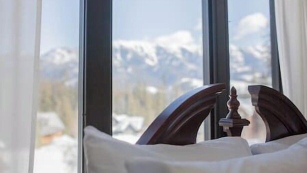 A bed with a wooden headboard and a view of snow-capped mountains through large windows.