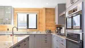 A modern kitchen with grey cabinets, a wooden wall, a large window, stainless steel appliances, and a granite countertop.