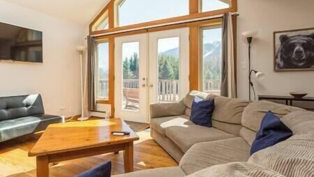 A spacious living room with a large wooden table, a grey sofa, and a balcony with a view of trees and mountains.