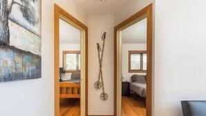 A hallway with wooden flooring, a painting on the left wall, and two beds with headboards in the background.