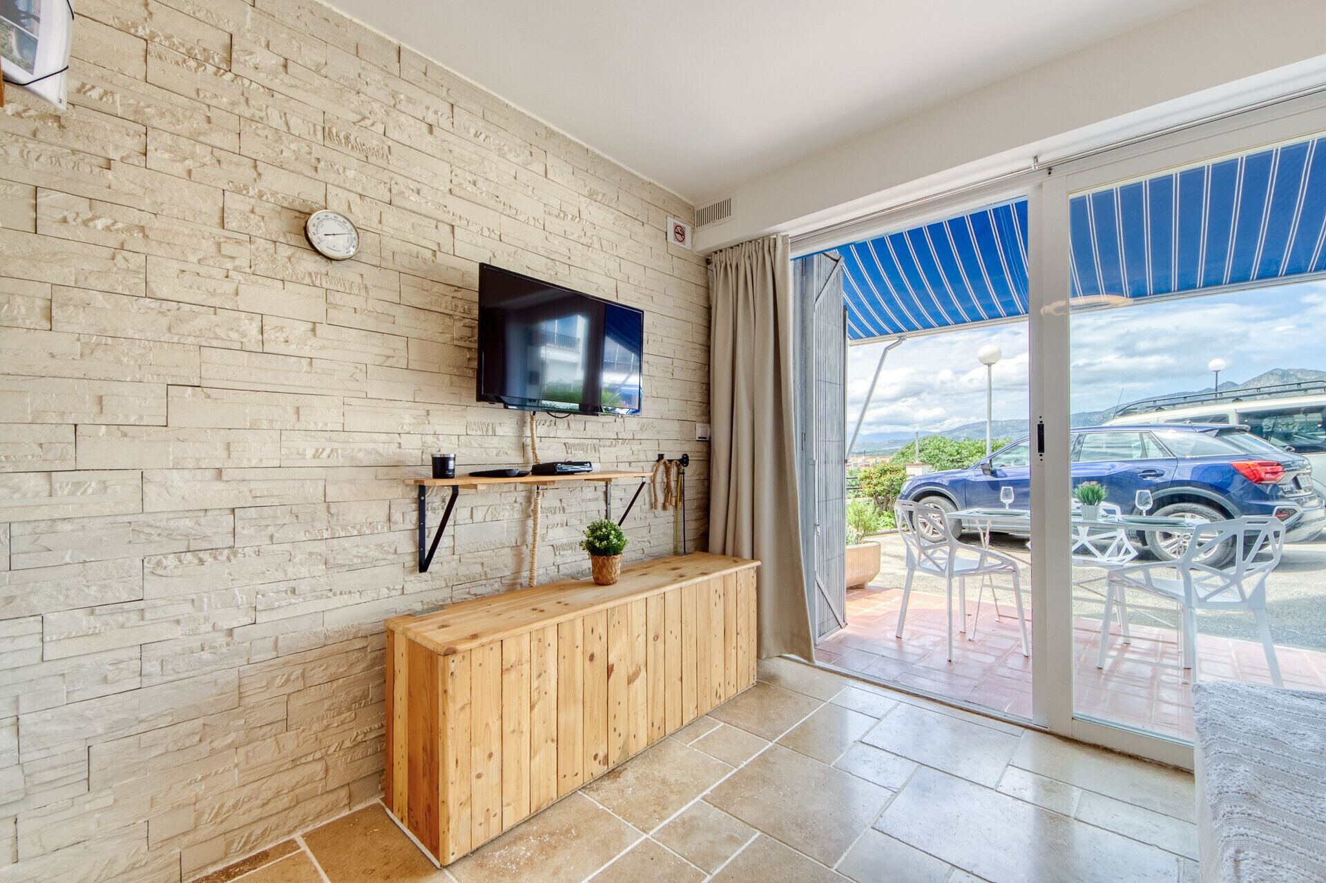 A modern living room with a brick wall, a flat-screen TV, a wooden bench, and a sliding glass door leading to an outdoor area with a car and a mountain view.
