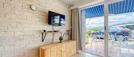 A modern living room with a brick wall, a flat-screen TV, a wooden bench, and a sliding glass door leading to an outdoor area with a car and a mountain view.