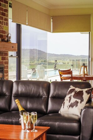 A modern living room with a leather sofa, a table with champagne glasses, and a view of a pool and hills. - Big Sky Country Views (Boyland)