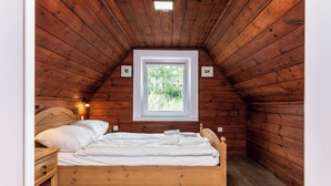 Ein Zimmer einer Holzhütte mit einem Einzelbett, einem Nachttisch, einem Fenster mit Blick auf Grünfläche und einer Deckenleuchte.