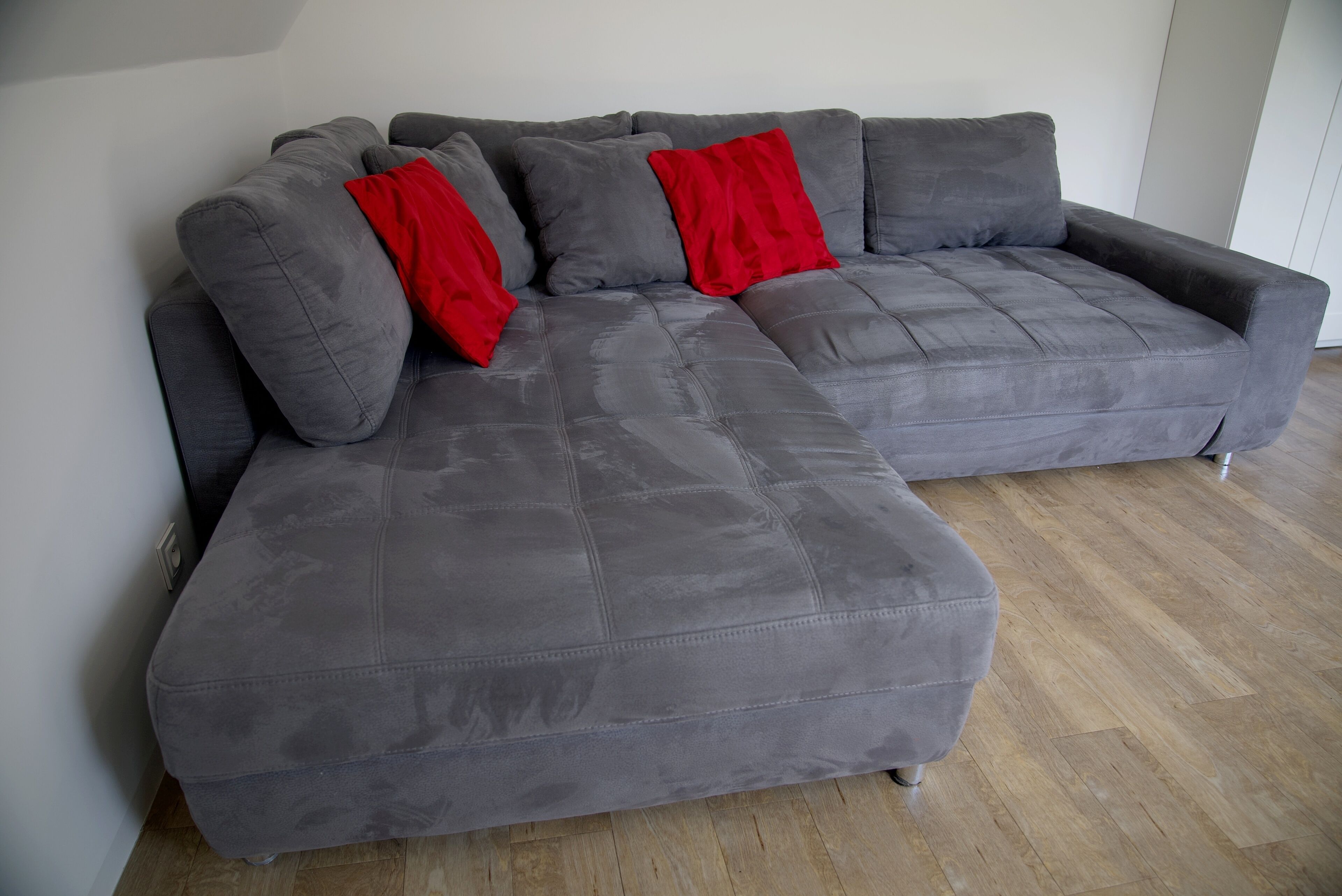 A grey sectional sofa with red cushions against a white wall, wooden flooring, and a power outlet on the wall.