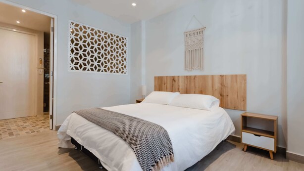 Une chambre à coucher moderne avec une tête de lit en bois, un lit recouvert d’une literie blanche, un plaid à motifs, une table de chevet en bois et une décoration murale en macramé.