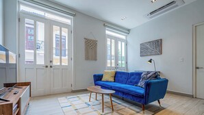 A living room with a blue sofa, a small wooden table, and a patterned rug. There are two windows with white frames and a ceiling fan. - w* | Airy 1BR w/ Balcony in Panama (Panamá)