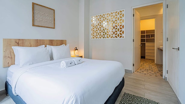 A neatly made bed with white linens and a rolled towel, a wooden headboard, a wall-mounted decorative panel, and a door leading to a tiled hallway. - w* | Airy 1BR w/ Balcony in Panama (Panamá)