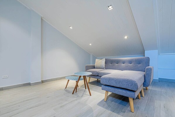 Un dormitorio estilo loft moderno con un sofá seccional gris, una mesita redonda y un techo inclinado con iluminación empotrada.