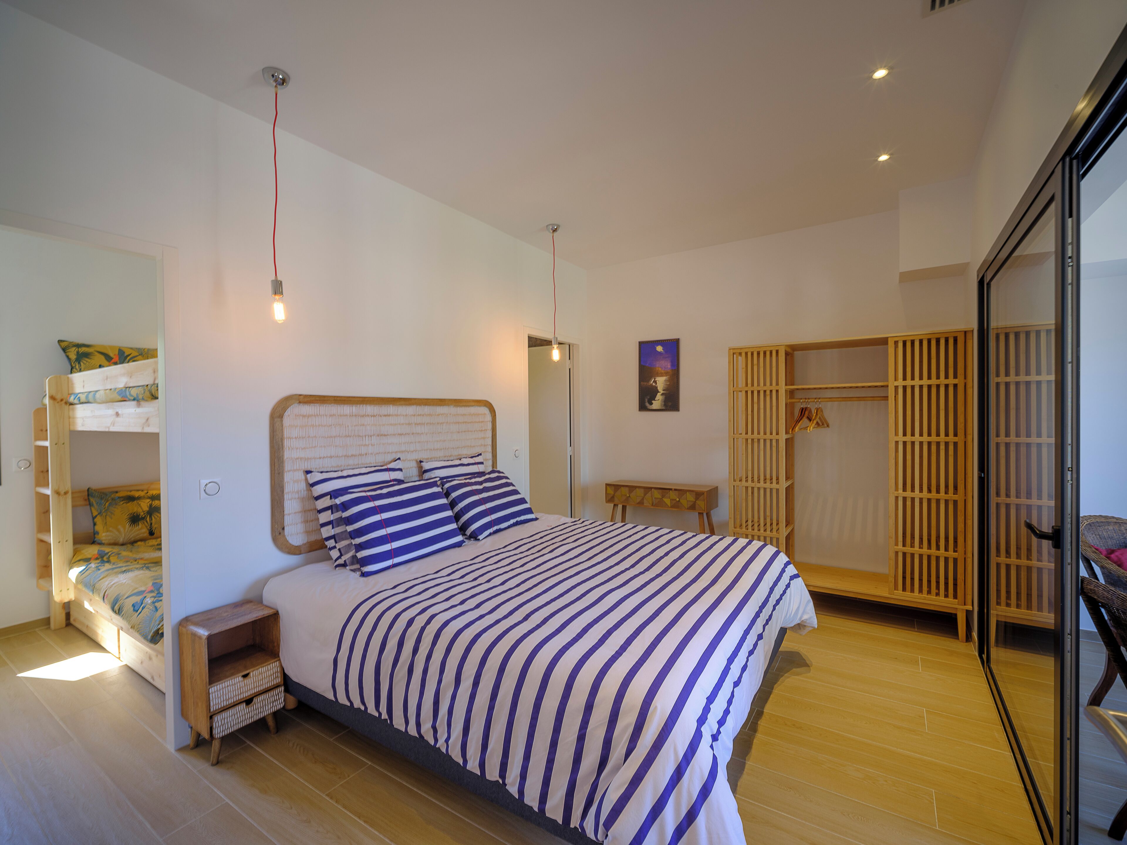 Un dormitorio con una litera, una cama grande con ropa de cama a rayas, una mesita de noche de madera y una pequeña mesa.