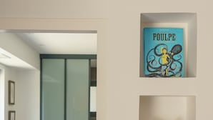 Huoneessa on sänky, tuoli ja seinälle kiinnitetty hylly, jonka päällä on kirja nimeltä ”POULPE” ja veistos.