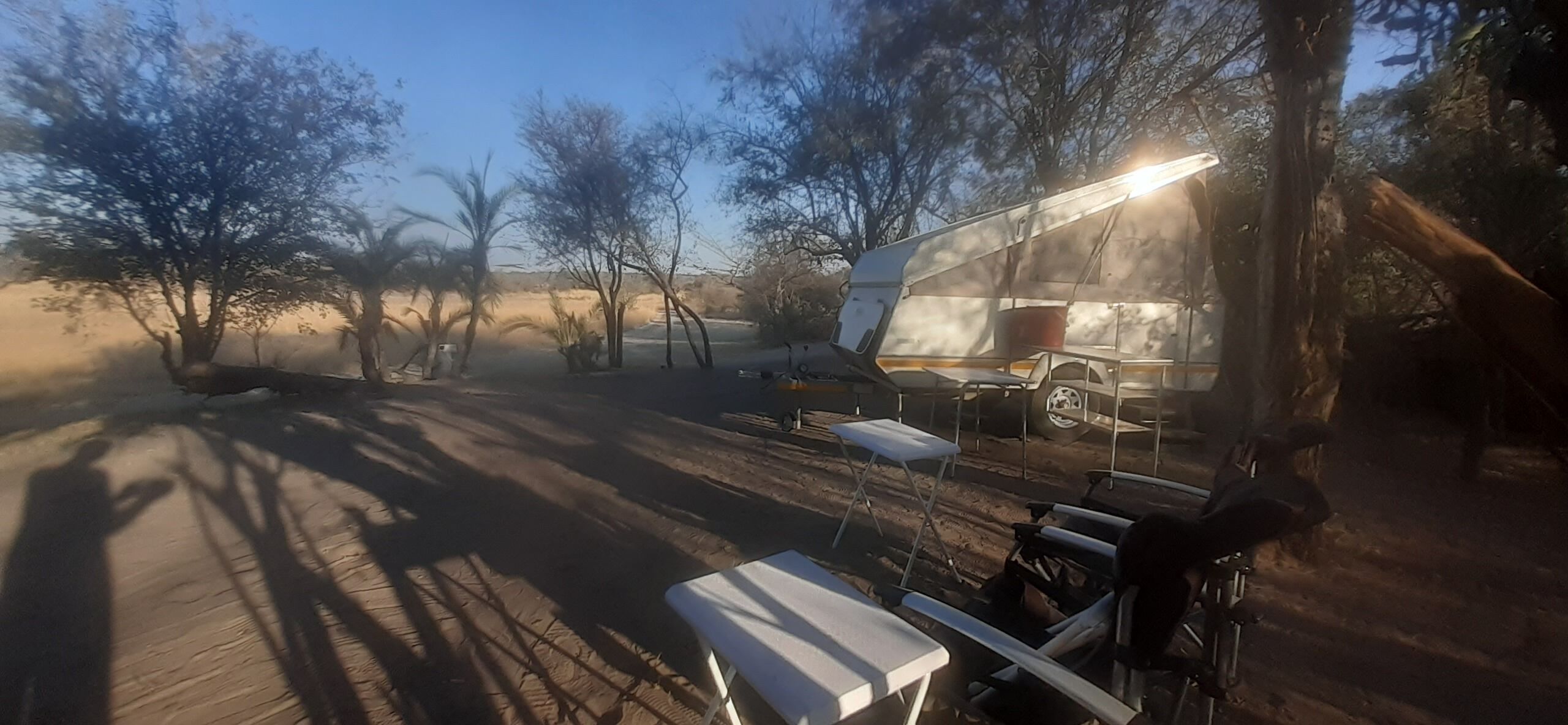 Un camping con una caravana grande, mesas y sillas dispuestas bajo los árboles.