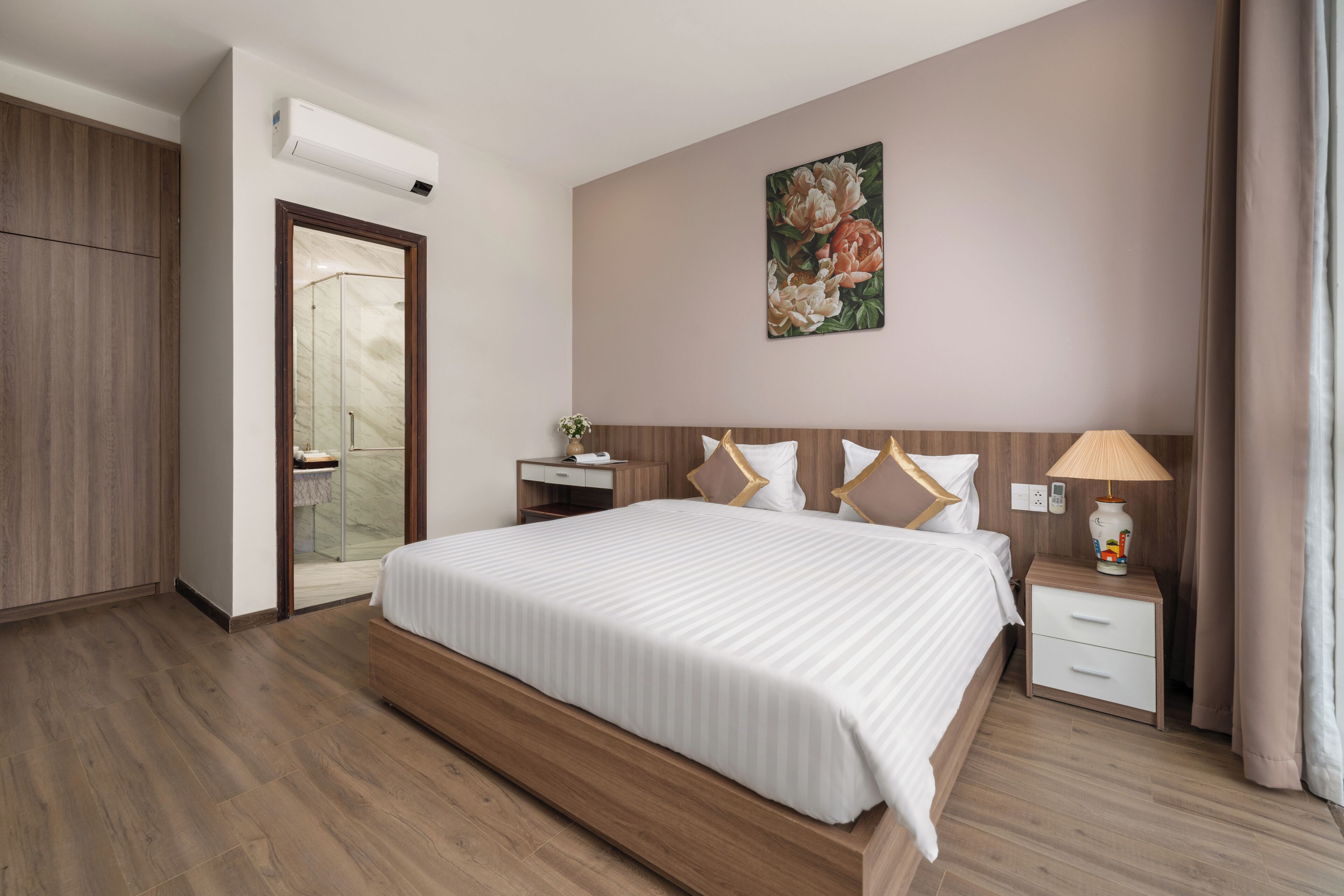 Habitación de hotel con una cama grande, suelo de madera, una mesita de noche con lámpara, un cuadro en la pared y una puerta que da a un baño.