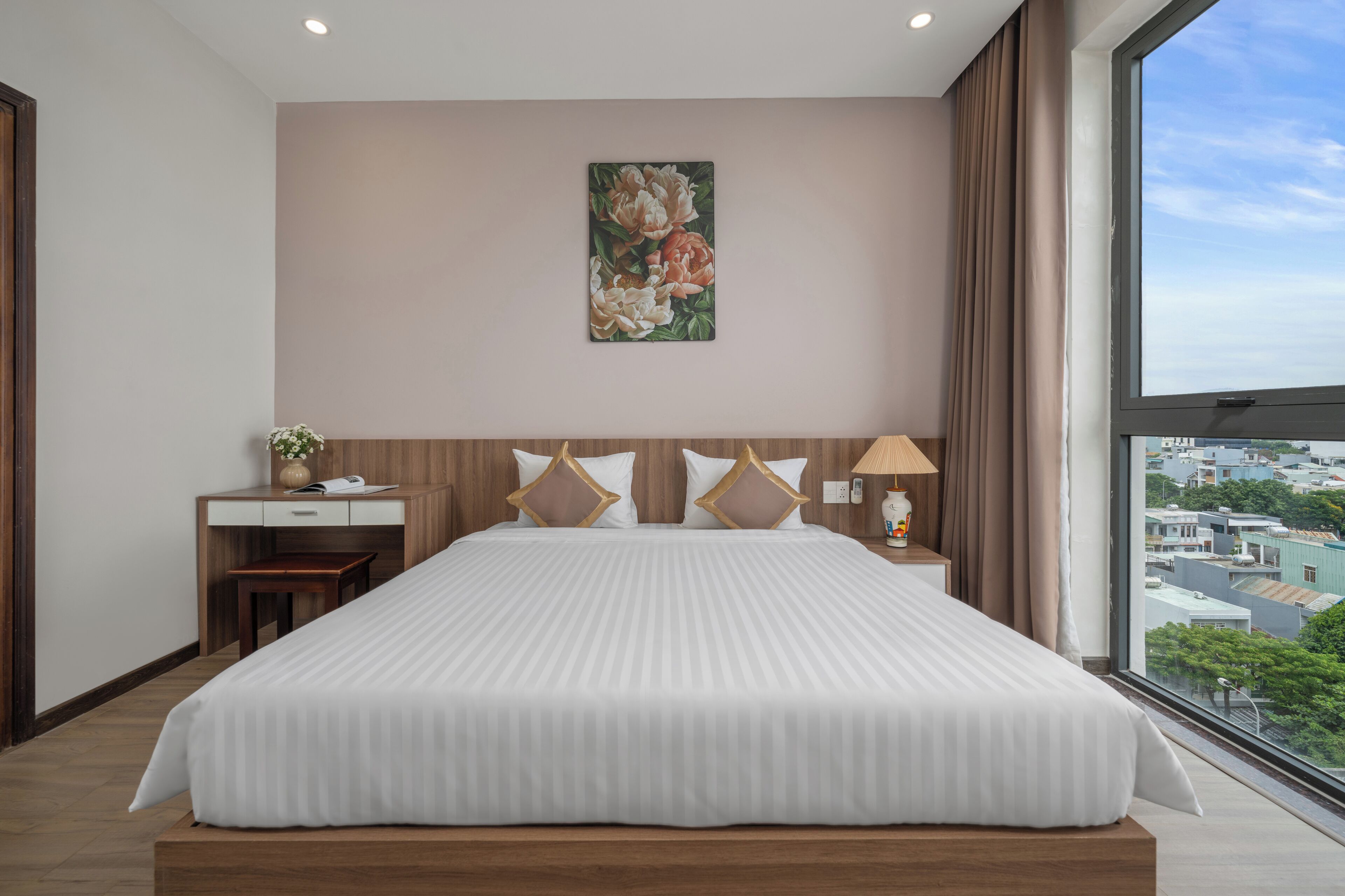 A modern hotel room with a large bed, a desk, and a view of the city through a large window.