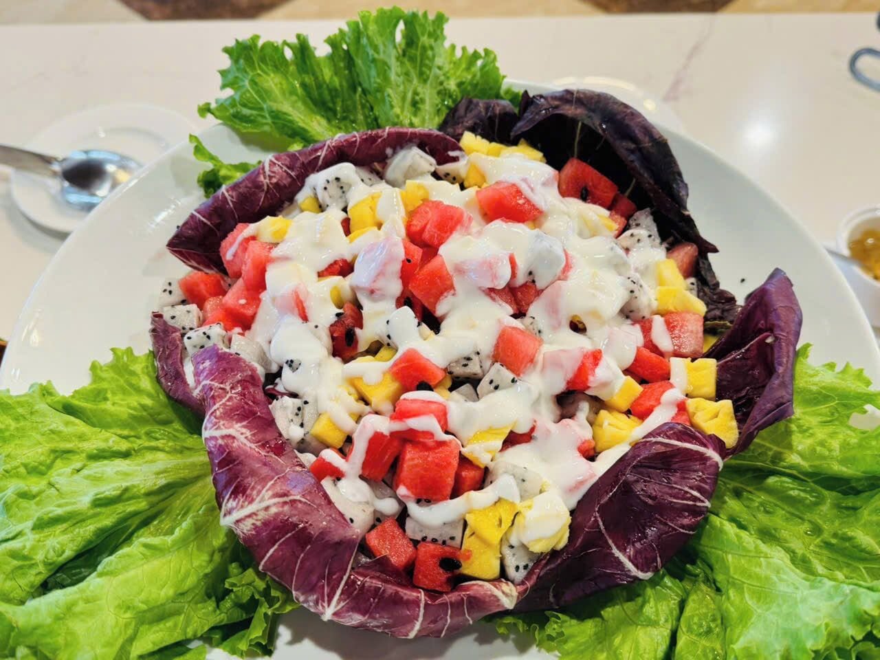 A salad with various fruits and a creamy dressing, served on a white plate with green lettuce leaves.
