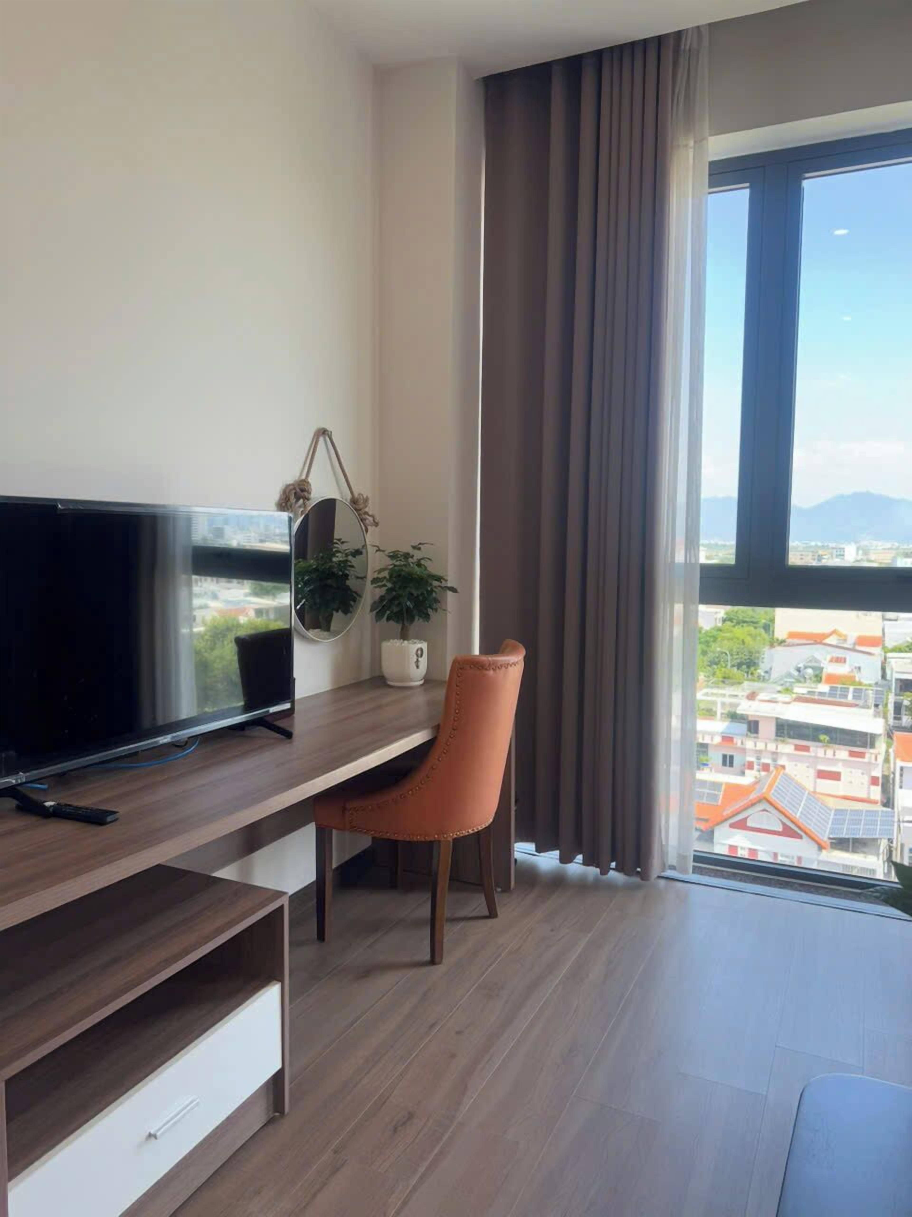 A modern hotel room with a wooden desk, a television, a chair, a potted plant, and a view of the city through large windows.
