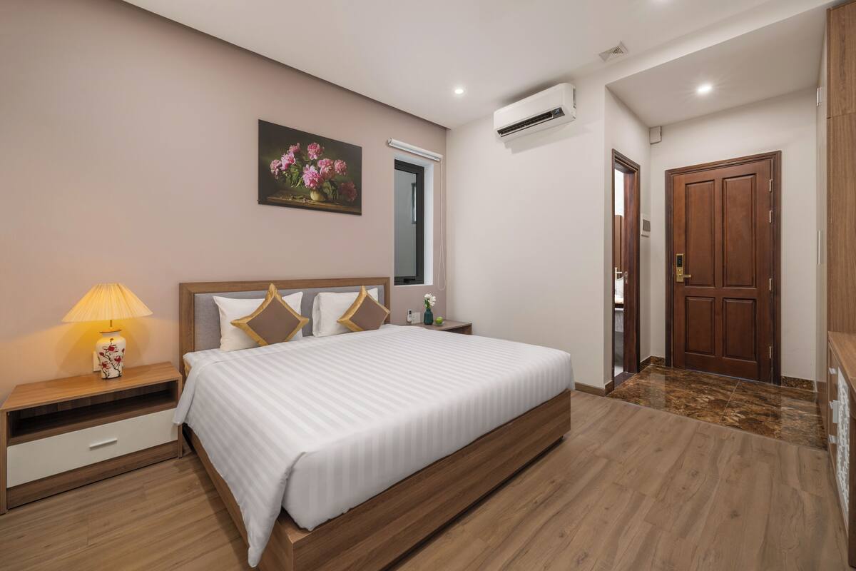 A bedroom with a large bed, a nightstand, a lamp, a painting, an air conditioning unit, and a wooden door.