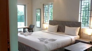 A modern hotel room with a large bed, a desk, and a chair. There are two windows with bars, a ceiling fan, and a nightstand with a lamp. - Hotel Peppers (Yercaud)