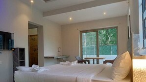 A hotel room with a bed, a desk, a chair, a small table, and a window with a view of greenery. - Hotel Peppers (Yercaud)