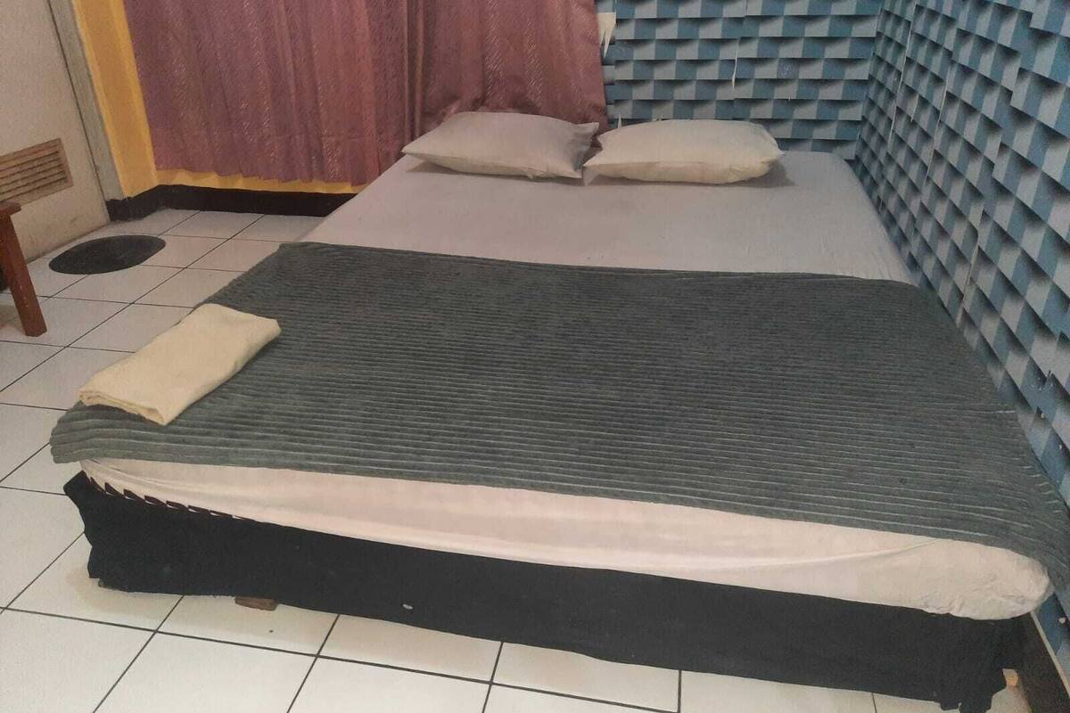 Ein Zimmer mit einem Bett, Kissen, einer Decke, einem hölzernen Hocker und einem Vorhang.