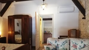A bedroom with a wooden bed, a floral sofa, a wooden wardrobe, and a kitchen visible through an open door.