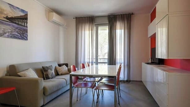 A modern living room with a dining table, sofa, and chairs. There is a painting on the wall, a window with curtains, and a kitchen area with cabinets. - Linda 19 - Lido di Jesolo, Italy. Entire Housing (Lido di Jesolo)