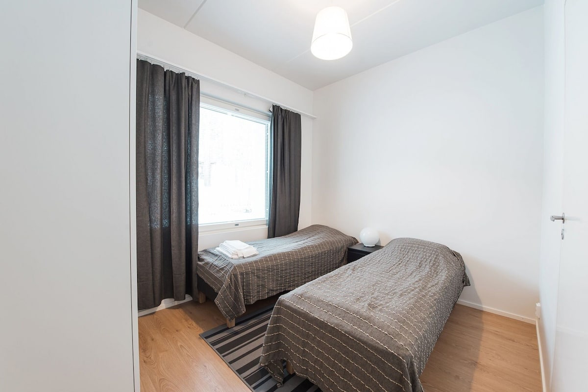 Een kleine, schone kamer met twee eenpersoonsbedden, een raam met gordijnen, een nachtkastje met een lamp en een houten vloer.